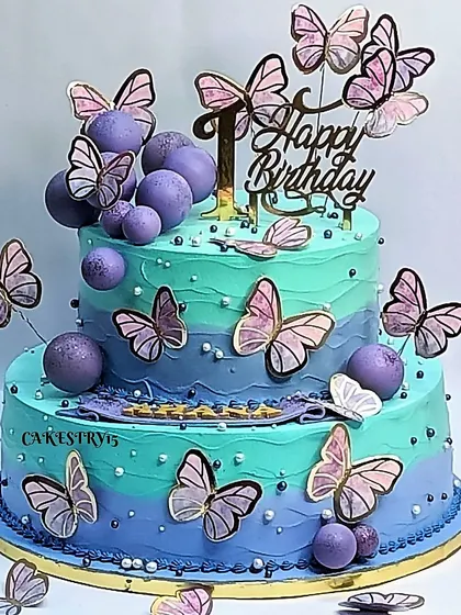 A stunning two-tier butterfly cake in shades of aqua and lavender. The cake is covered in beautiful paper butterflies and decorative purple balls for a vibrant and magical look.