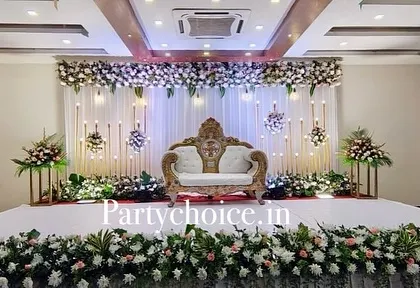 A grand and elegant engagement stage featuring a white floral header, a draped backdrop with candle-like lights, and a traditional royal sofa.