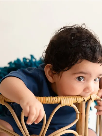 A curious baby in a blue outfit peeks over the side of a wicker basket during his sitter session.