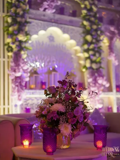 A detail shot of a side table, featuring a beautiful floral arrangement and purple glass candle holders. The intricate white backdrop, lit with purple light, creates a stunning setting.
