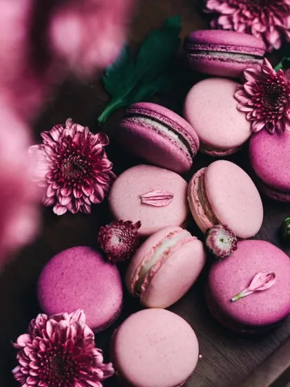 A celebration of pink with these beautiful macarons. The moody, dark lighting contrasts with the delicate pink hues and fresh flowers, creating a romantic and artistic dessert photograph.