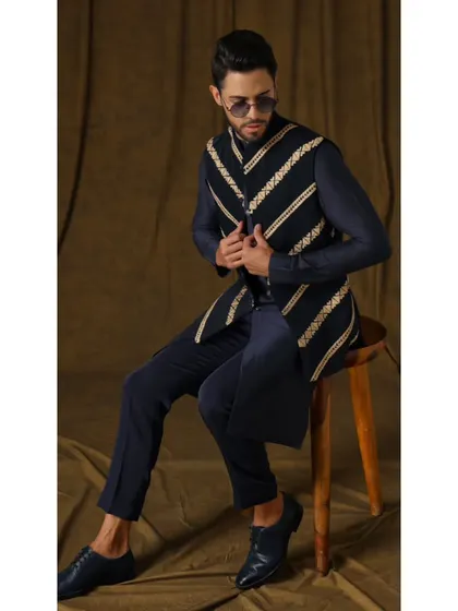 A seated pose in the 'Nilo Pushpak' ensemble. This view offers a different perspective on the jacket's wired embroidery and the sleek fit of the cigarette trousers.