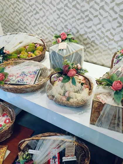 A curated collection of trousseau gifts showcasing different packing styles. We use wicker baskets for fruits, geometric glass boxes for delicate items, and fabric-wrapped gifts, all unified with soft pink floral accents.