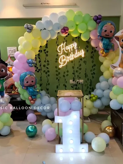 This Cocomelon first birthday setup for a home party includes a pastel balloon arch over a greenery wall, a neon sign, character cutouts, and a light-up number '1'.