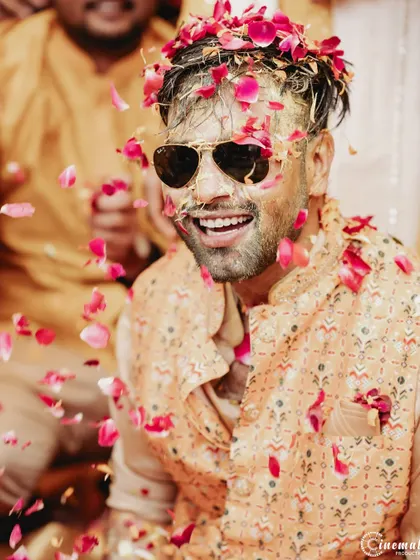 The groom, wearing sunglasses and covered in haldi and petals, laughs for the camera. A perfect shot of the fun-filled Haldi madness.