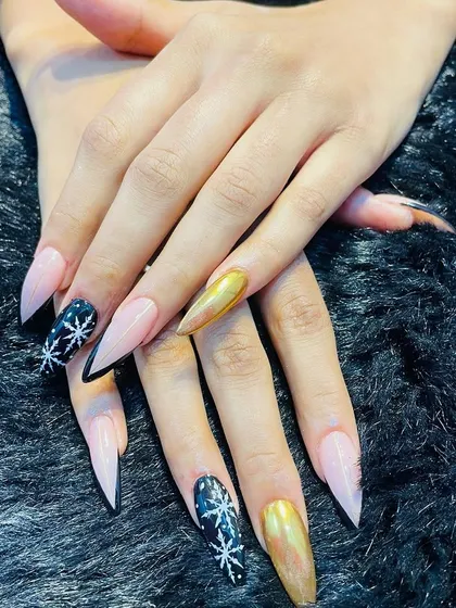 A dramatic and festive set of long stiletto nails. The design combines a sheer pink base with black nails featuring sparkling snowflakes and a stunning gold chrome accent nail.