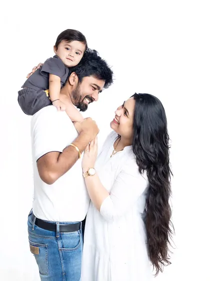 A sweet family portrait against a clean white background. The parents look at each other while their baby sits on dad's shoulders, creating a lovely, connected image.