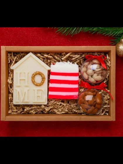 A cozy Christmas-themed gift set. This hamper includes a "Home" soy wax candle, a festive striped soap, and dry fruits, all nestled in a wooden tray.