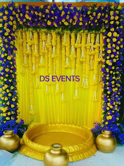 A striking color combination for a Haldi ceremony. This backdrop uses a frame of deep blue and bright yellow flowers, with hanging marigold and jasmine garlands over a yellow silk curtain.