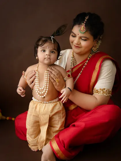 A close-up portrait of a mother admiring her little Krishna. The simple brown background puts all the focus on their connection.