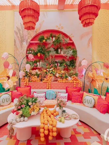 This vibrant seating lounge features a custom-built backdrop with tiered shelves of florals and fresh oranges. We used playful 'Oui' cushions and bold red and orange textiles on clean white sofas to create a fun, high-contrast look.