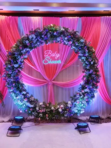 A stunning circular floral arch set against a backdrop of pink and white drapery. The 'Baby Shower' neon sign in the center adds a modern touch to this grand and beautiful stage decoration.