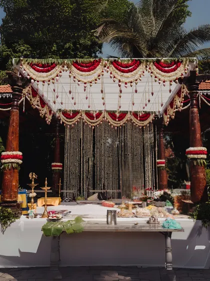A classic South Indian mandap setup at The Peacock Grove, featuring traditional wooden pillars and a canopy decorated with red and white floral scallops and hanging tuberose strings. This design honors heritage with timeless elegance.