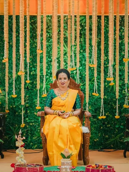 A bride seated against a stunning backdrop of green foliage and hanging marigold strings for her traditional Haldi ceremony.