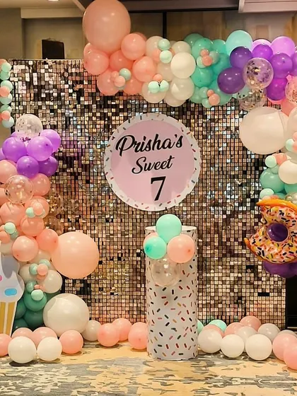 A "Sweet 7" birthday party with a donut and cupcake theme. The setup includes a shimmer wall, a pastel balloon arch, and large inflatable donut and cupcake props.