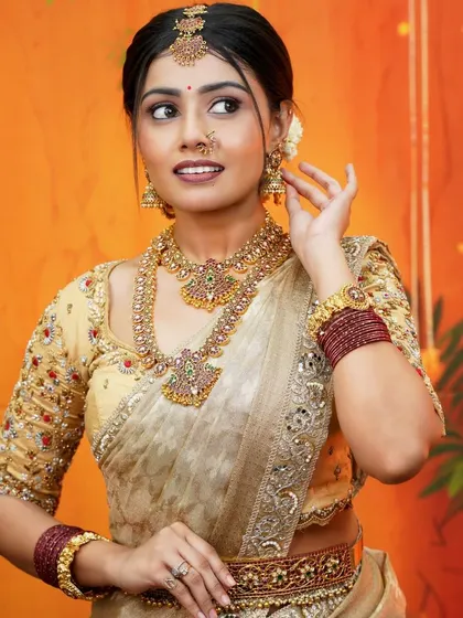 A beautiful portrait of a bride in a cream and gold silk saree, showcasing the elegance of a Muhurtham look. The warm, festive background highlights the saree's golden sheen.