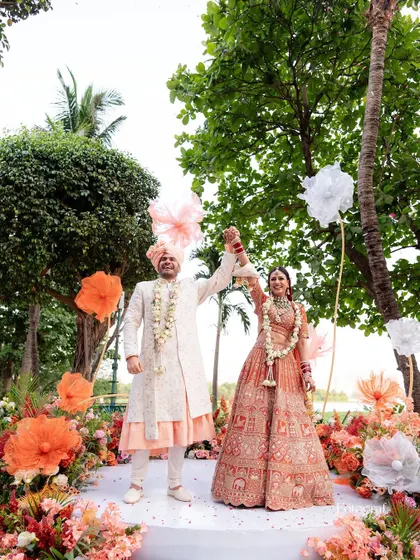 The newlyweds celebrating with hands raised high after their Varmala ceremony, surrounded by beautiful floral decor.