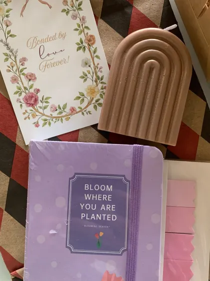 This minimal Rakhi gift set features an arch-shaped scented candle and a lavender journal with the quote 'Bloom where you are planted,' perfect for a sister who loves stationery.