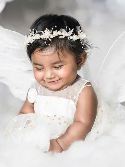 A baby girl dressed as an angel, looking down with a shy, sweet smile. The soft lighting and cloud-like setting create a heavenly feel.