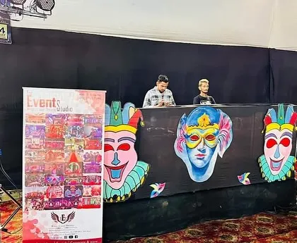 The DJ console for a carnival themed event, featuring a custom designed facade with colorful masks. Our branding standee is also visible, showcasing our professional presence.