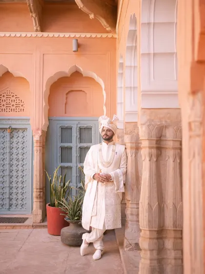 A solo portrait of the groom in his white sherwani, looking regal as he leans against a pillar in a sunlit palace courtyard.