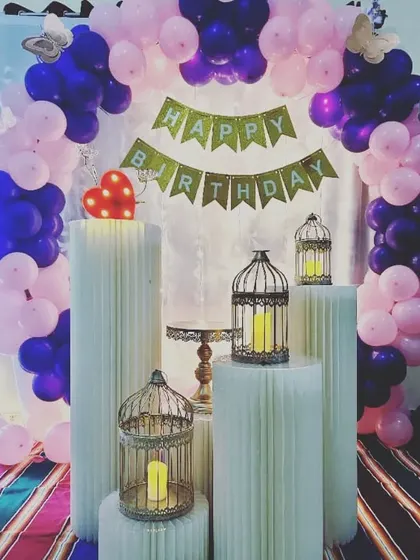 A simple home birthday decoration with a purple and pink balloon arch, a 'Happy Birthday' banner, and decorative birdcages.