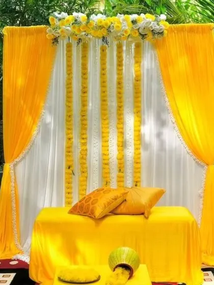 An outdoor Haldi setup using the natural greenery as a background. The decor features a simple white drape with hanging marigold strings and a seating arrangement covered in bright yellow fabric.