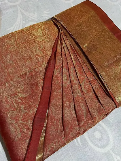 A beautiful rust-colored silk saree with intricate gold work. The pleats are set to ensure the saree falls gracefully.