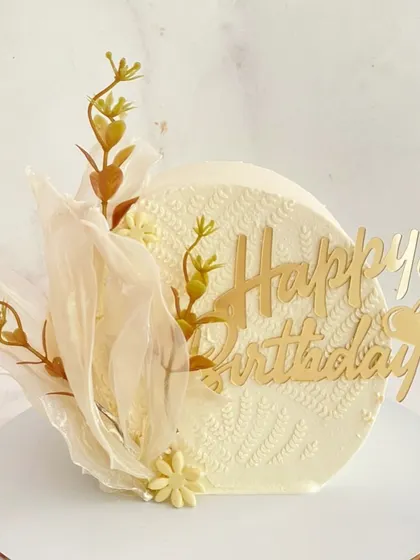A minimalist and artistic white birthday cake. It features a textured, knitted pattern on the surface, a delicate rice paper sail, and a gold acrylic "Happy Birthday" topper.