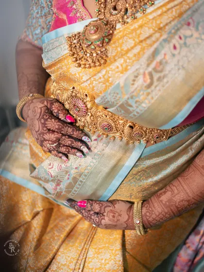 A close-up on the details: the mother's hands with intricate henna, the rich texture of the saree, and the ornate gold waist belt (vaddanam).