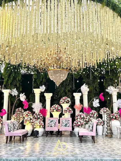 A majestic floral garden stage, featuring a massive chandelier made of tuberoses hanging over a setting of pink and white. The Roman pillars add a touch of classic grandeur.