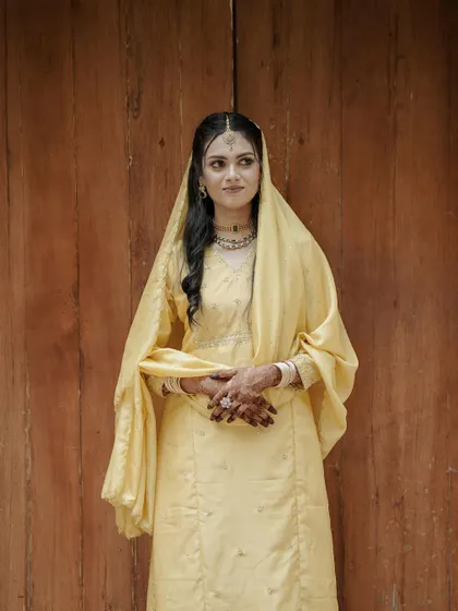 A full-length portrait of the bride against a simple wooden background. This minimalist approach puts all the focus on her elegant yellow salwar suit and her graceful posture.