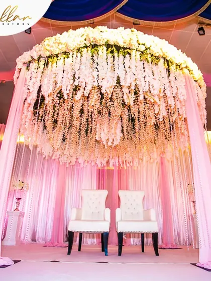 A stunning mandap or stage design with a canopy made entirely of hanging white flowers. The soft pink lighting creates a dreamy and ethereal ambiance.