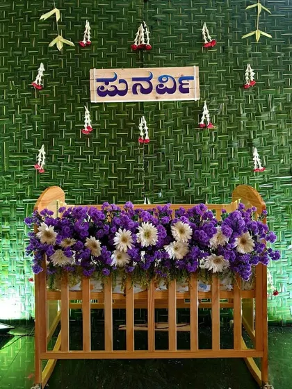 A naming ceremony setup with a wooden cradle, purple flowers, and a woven coconut leaf backdrop featuring a name in Kannada.