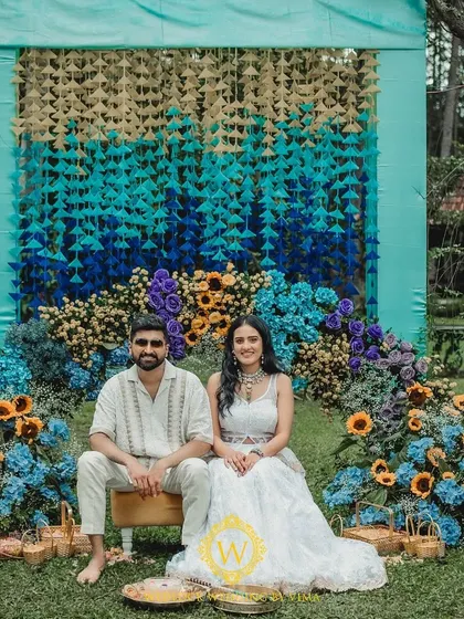 A beautiful setup for a vibrant Haldi and Mehendi ceremony. The backdrop features a stunning gradient of hanging tassels in shades of blue and yellow, complemented by a floral arch of sunflowers and hydrangeas.