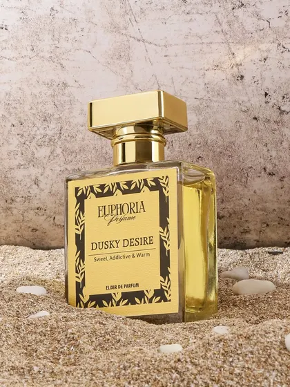 A beach inspired shot for 'Dusky Desire' perfume, placing the bottle in the sand. This setting evokes a sense of warmth, holidays, and sensuality, matching the 'Sweet, Addictive & Warm' description.