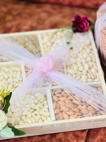 A close-up of a beautifully arranged dry fruit gift box, part of a larger trousseau. The wooden box with separate compartments for cashews, almonds, and pistachios is wrapped simply with net to showcase the contents.