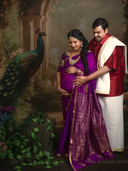 An expecting couple stands in a regal, vintage-inspired studio setting with a painted peacock backdrop. This traditional maternity portrait is rich with cultural elegance and artistry.