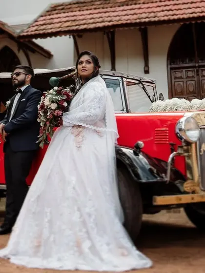 A couple in their wedding attire posing with a classic red Austin car outside a church. This image combines timeless elegance with a touch of vintage charm.