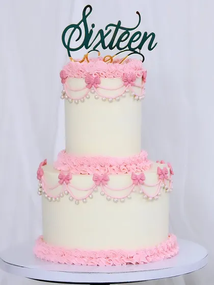 A classic two-tiered cake for a sweet sixteen celebration. This design features intricate, vintage-style pink piping and pearl details against a clean white buttercream base.
