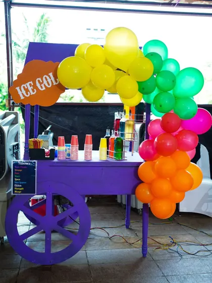 A vibrant purple ice gola cart decorated with a rainbow balloon arch, perfect for cooling down at a fun-filled Punjabi-themed party.