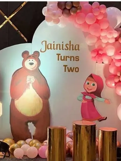 A sweet Masha and the Bear theme for a second birthday. The setup includes a custom backdrop with the characters, a pink and gold balloon arch, and a light-up number '2'.