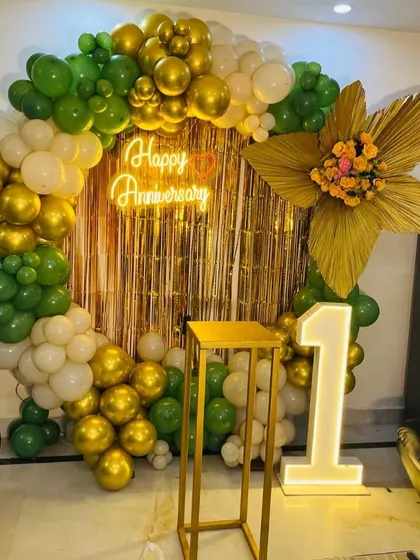 A beautiful anniversary backdrop with a balloon ring in green, white, and gold, a neon sign, and a large floral accent made of golden leaves.
