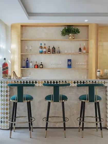 This bespoke home bar is where gold accents meet velvet green bar chairs. The counter features a unique diamond-patterned inlay, and the open shelving is lit to showcase the collection, creating an inviting corner for a pour and a pause.