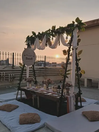 A beautiful sunset picnic setup on a terrace. The low table is set for a party, with a decorative sign and a frame adorned with greenery.