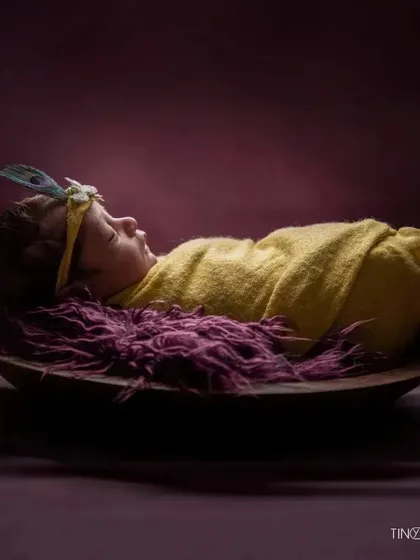 A simple yet striking portrait of a newborn with a peacock feather headband. The baby is swaddled in yellow and rests on a rich purple fur, creating a beautiful color contrast.