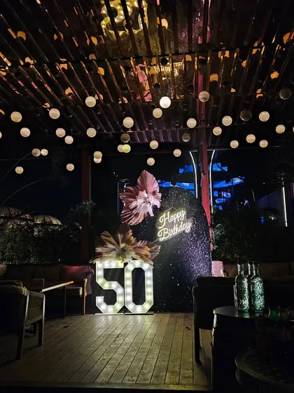 Celebrating a 50th birthday calls for something truly special. For this milestone party, I designed a sophisticated outdoor lounge setting with a dramatic sequin backdrop, a custom neon sign, and large marquee numbers, all under a canopy of warm string lights.