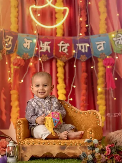 A smiling baby boy celebrating Diwali, seated on a small couch against a backdrop of marigold flowers and lights. The neon "diya" adds a modern touch.