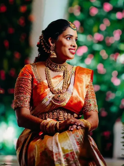 A beautiful portrait of a bride in a rich orange Kanjeevaram saree with a heavily embroidered blouse, looking radiant against a floral backdrop.