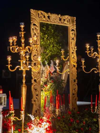 An ornate, oversized gold mirror reflects the candlelight and red florals, adding depth and glamour to the decor. The candelabras in front enhance the feeling of old-world opulence.